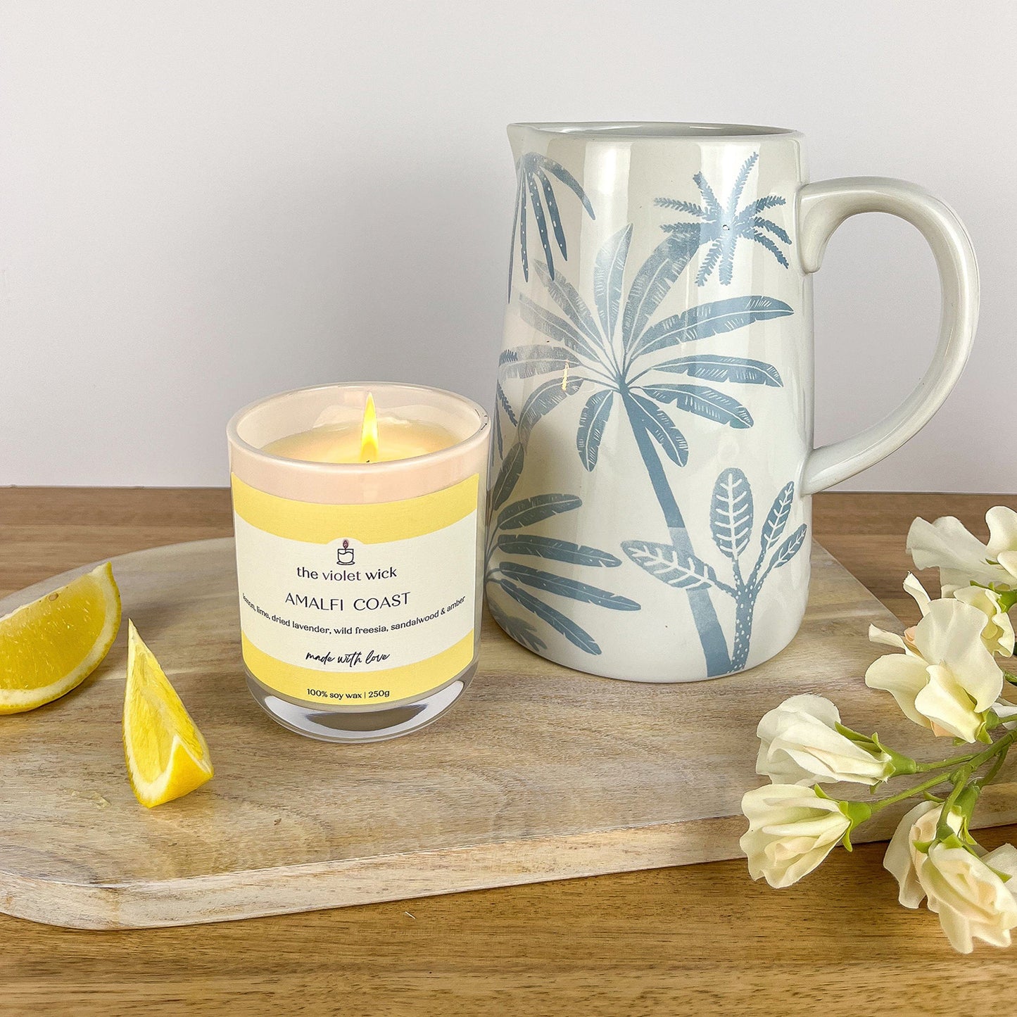 Amalfi Coast scented soy candle from The Violet Wick on a serving tray with lemon slices, a jug and flowers. In a glass jar with timber lid, 250g.