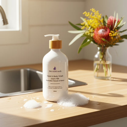 Australian Florals & Honey Goat's milk hand and body wash bottle on light timber kitchen bench with Australian flowers in a vase in background.