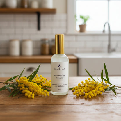 Bottle of 'The Violet Wick' Bush Blossom Room Spray on a timber kitchen bench with golden bush blossoms