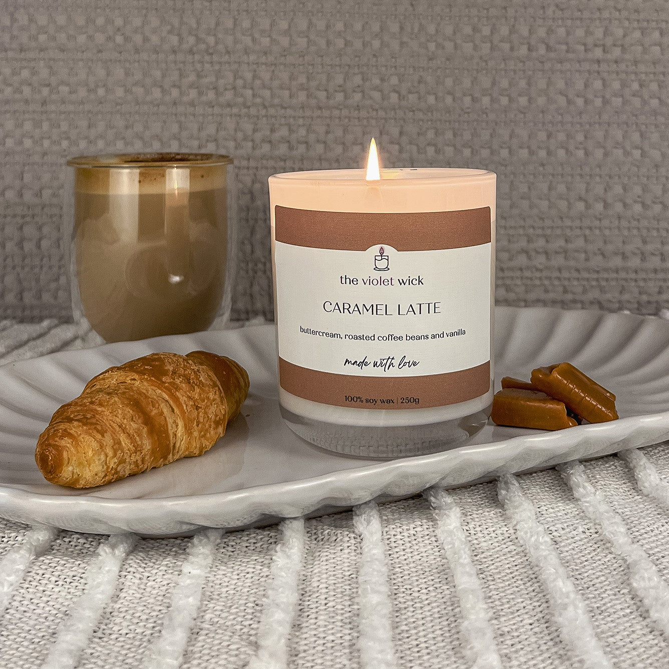 Caramel Latte scented soy candle from The Violet Wick in glass jar with timber lid, 250g. Served on a dish with latte, croissant and caramel lollies. 