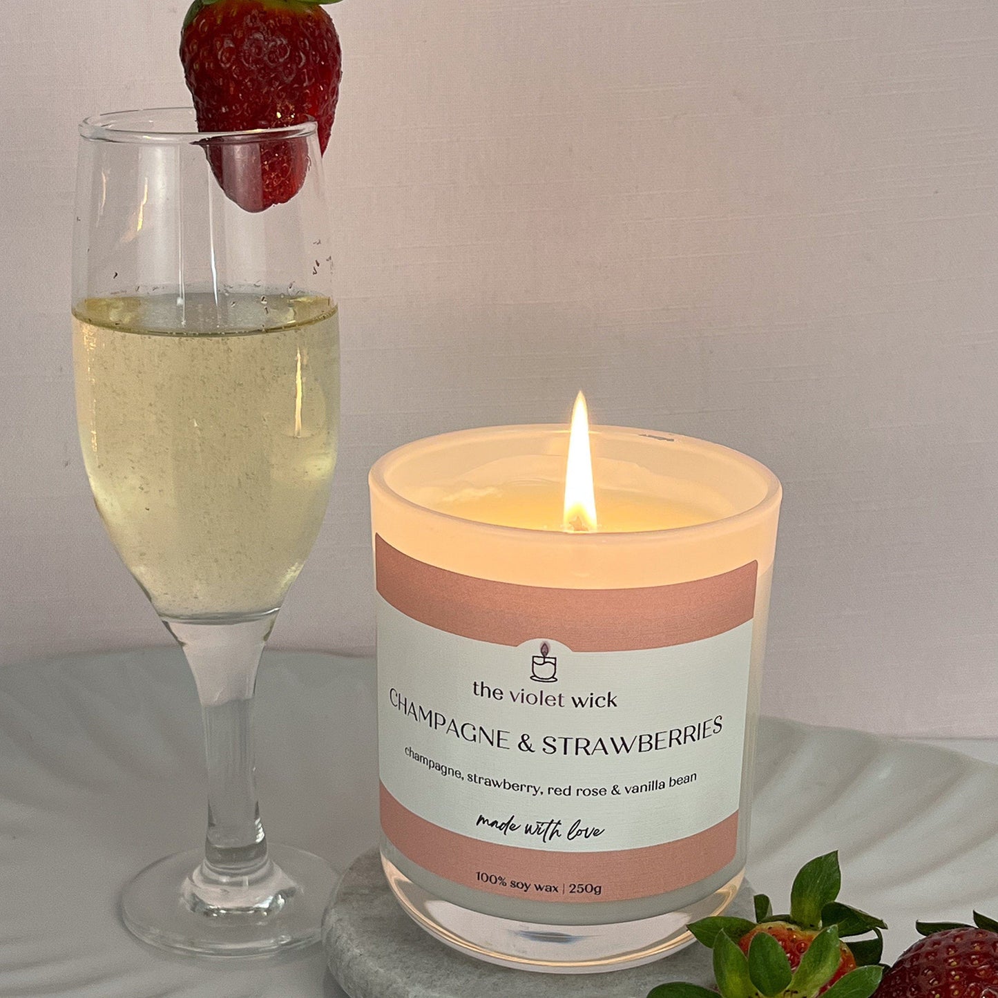 Champagne &. Strawberries scented soy candle from The Violet Wick in a glass jar in a serving plate with glass of champagne surrounded by strawberries, 250g.