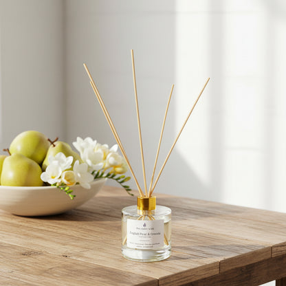 clear reed diffuser bottle with gold lid labeled English Pear & Freesia on coffee table with a bowl of pears and freesia