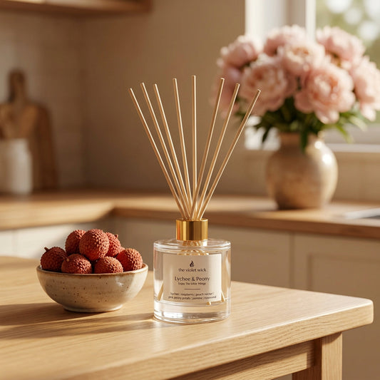 Lychee & Peony Reed Diffuser by The Violet Wick on kitchen bench with a bowl of lychees and vase of peonies in background.