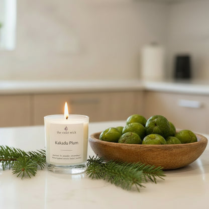 Candle labeled 'Kakadu Plum' by 'the violet wick' on a kitchen bench with kakadu plums in a bowl and fir needle.