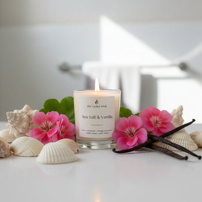 Candle labeled 'Sea Salt & Vanilla' by 'the violet wick' on a white bathroom bench with sea shells, geraniums and vanilla pods.